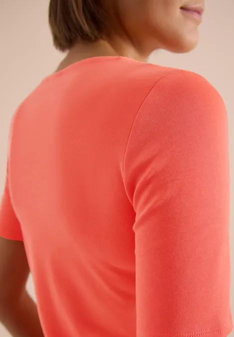 Rugaanzicht van een persoon in een koraalkleurig T-shirt met korte mouwen tegen een lichte achtergrond.