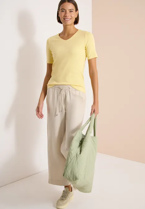 Basic katoenen shirt buttermilk yellow Vrouw in geel T-shirt en beige wijde broek, met groene tas en witte sneakers.
