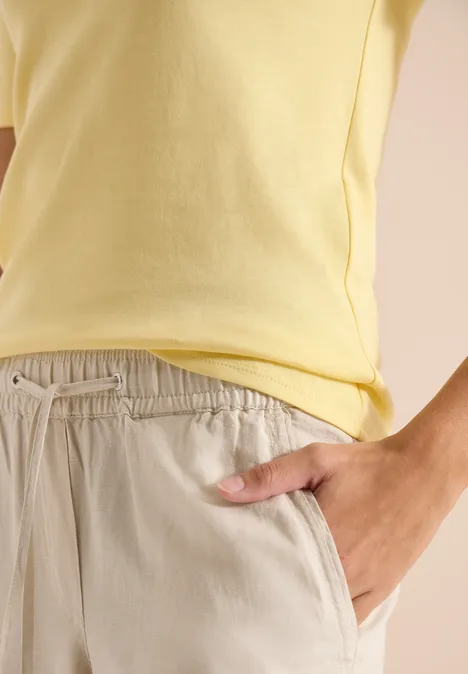 Basic katoenen shirt buttermilk yellow Close-up van een lichtgeel T-shirt en beige broek, hand in de zak.