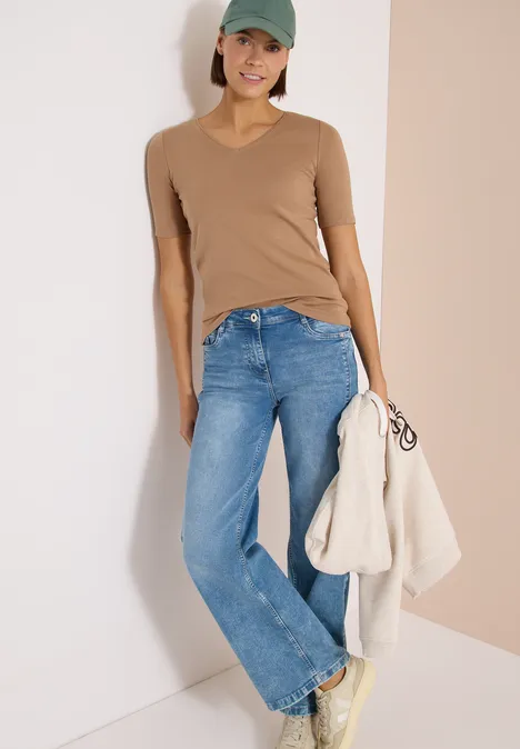 T-shirt basique en coton creamy mocha Femme appuyée contre un mur, porte un t-shirt col V beige, jean bleu et tient une veste claire.