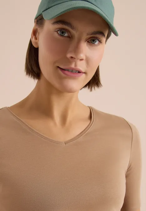 T-shirt basique en coton creamy mocha Personne avec casquette verte et t-shirt beige à col en V sur fond clair, visage flouté.