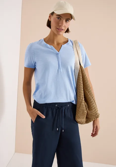 Vrouw draagt lichtblauw T-shirt, donkerblauwe broek, pet en gevlochten strozak voor neutrale achtergrond.