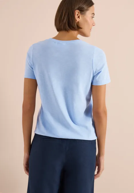 Rugzicht van een persoon in lichtblauw T-shirt en donkerblauwe broek tegen een beige achtergrond.