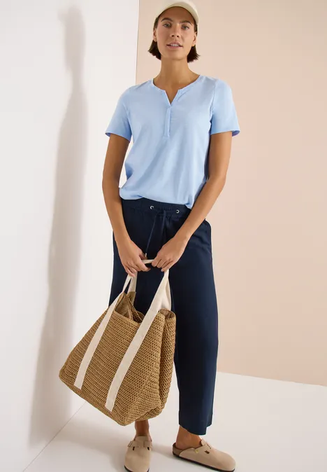 Vrouw in lichtblauw topje en donkerblauwe broek, met grote rieten tas en beige slippers voor een lichte muur.