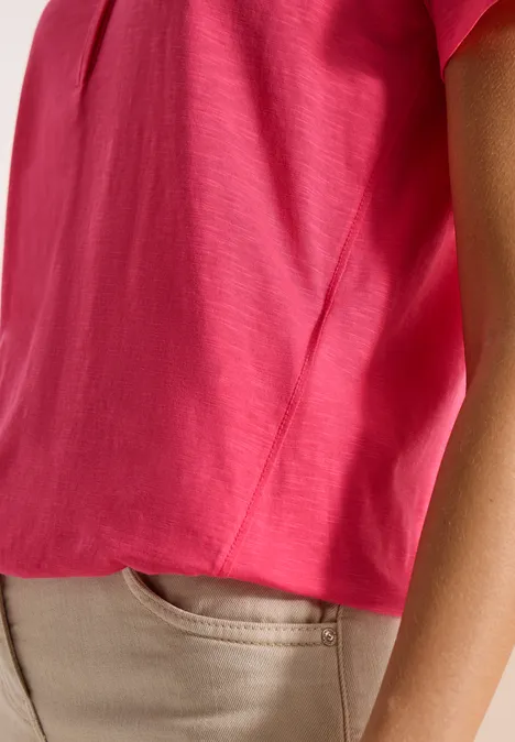 Close-up van een persoon met een felroze top en een beige broek, alleen torso en arm zichtbaar.