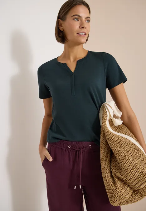 Frau in dunklem T-Shirt und weinroter Hose, trägt eine große, geflochtene Strohtasche.
