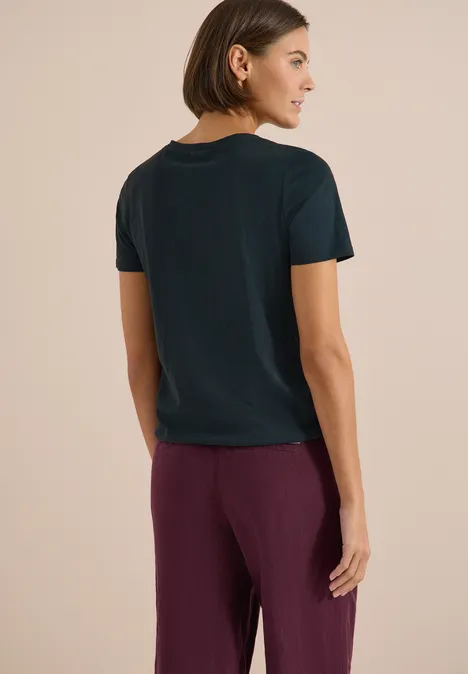 Person mit schulterlangem Haar, dunklem T-Shirt und lila Hose, stehend vor beigem Hintergrund.
