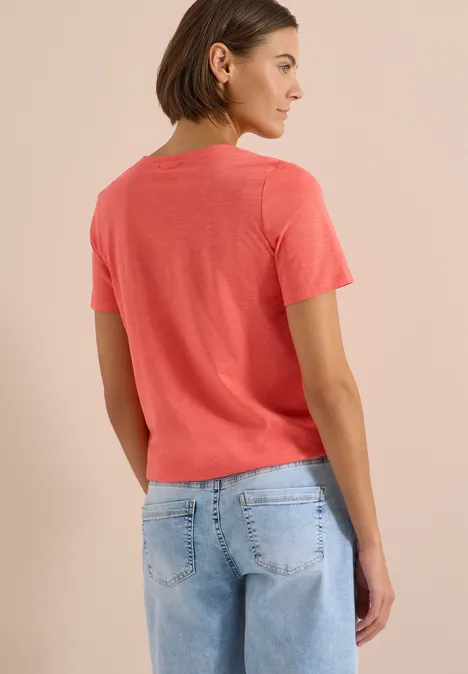 Frau steht von hinten, trägt ein korallenrotes T-Shirt und blaue Jeans vor neutralem Hintergrund.