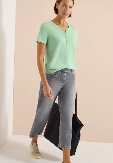 Basic shirt in tuniekstijl juicy lime Vrouw met mintgroene blouse, grijze jeans, witte sneakers en een zwarte tas tegen een beige achtergrond.