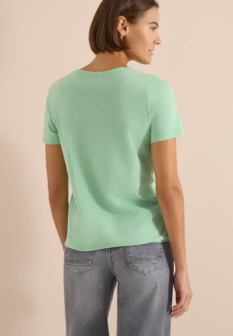 Basic shirt in tuniekstijl juicy lime Persoon van achteren in mintgroen T-shirt en lichte spijkerbroek voor een beige achtergrond.