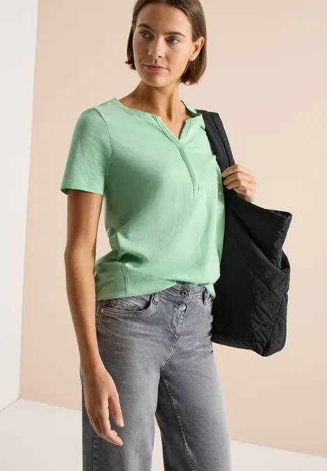 Basic shirt in tuniekstijl juicy lime Vrouw met groen poloshirt, grijze jeans en zwarte tas tegen neutrale achtergrond.