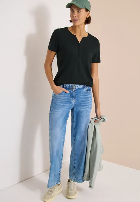 Basic shirt in tuniekstijl palm breeze green Persoon draagt jeans, zwart T-shirt en pet, houdt een jas vast, staat voor een lichte achtergrond.