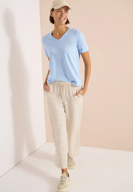 Basic Shirt met V-hals light sky blue Persoon draagt lichtblauw T-shirt, beige linnen broek en sneakers, staand ontspannen in een lichte ruimtehoek.