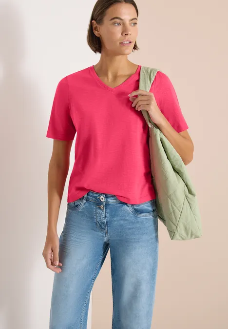 Basic Shirt met V-hals geranium red Vrouw in roze T-shirt, spijkerbroek en groene doorgestikte schoudertas voor neutrale achtergrond.