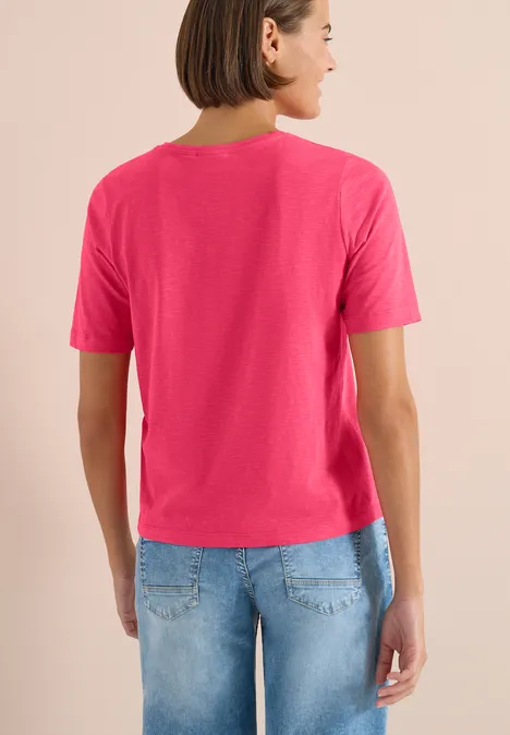 Basic Shirt met V-hals geranium red Een vrouw van achteren draagt een felroze T-shirt en lichtblauwe spijkerbroek.