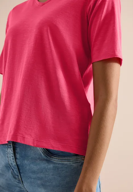 Basic Shirt met V-hals geranium red Bovenlichaam in een roze V-hals T-shirt en blauwe spijkerbroek, zichtbaar van schouder tot heup.