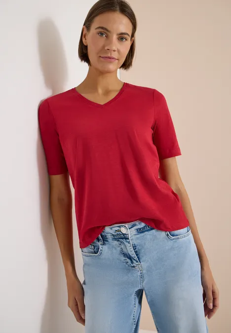 Basic T-Shirt mit Kurzarm und V-Neck tamarillo red Eine Person in rotem T-Shirt mit V-Ausschnitt und hellen Jeans lehnt vor einer hellen Wand.