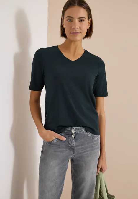 Frau trägt dunkles T-Shirt und graue Jeans, hält grünes Tuch in der Hand vor weißem Hintergrund.
