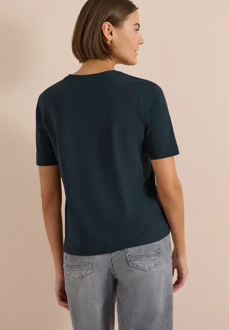 Frau von hinten in marineblauem T-Shirt und grauer Jeans vor beigem Hintergrund.