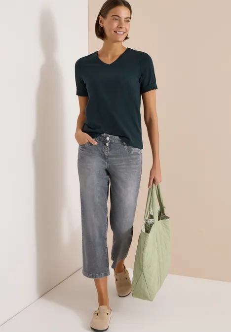 Frau in schwarzem T-Shirt, weiten Jeans-Culottes und hellen Schuhen, trägt eine grüne Tragetasche.