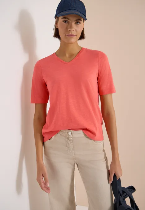 Basic T-Shirt mit Kurzarm und V-Neck shiny coral Person mit rotem V-Shirt, beigen Hosen und blauer Mütze, hält eine dunkelblaue Tasche.