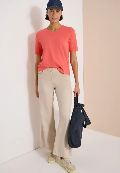 Basic T-Shirt mit Kurzarm und V-Neck shiny coral Person vor Wand in korallfarbenem T-Shirt, heller weiter Hose und Sneakers, hält Tasche.