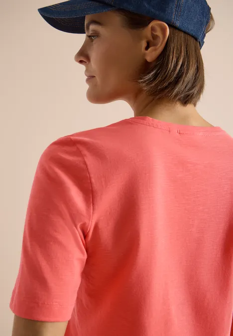 Basic T-Shirt mit Kurzarm und V-Neck shiny coral Rückenansicht einer Person mit korallfarbenem T-Shirt und blauer Kappe vor beigem Hintergrund.