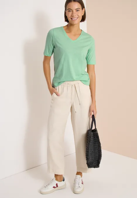 Basic Shirt met V-hals juicy lime Persoon voor muur, draagt lichtgroen V-shirt, losse beige broek, witte sneakers en zwarte tas.