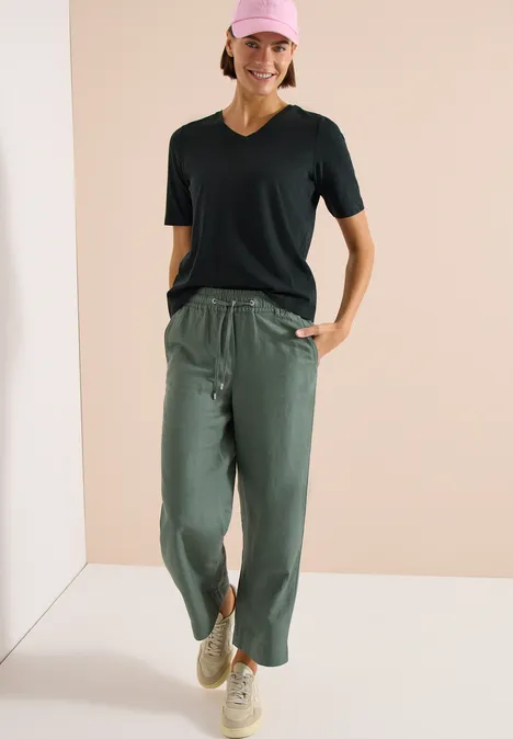 Basic Shirt met V-hals palm breeze green Vrouw in een zwart T-shirt, losse olijfgroene broek met tunnelkoord en beige sneakers.