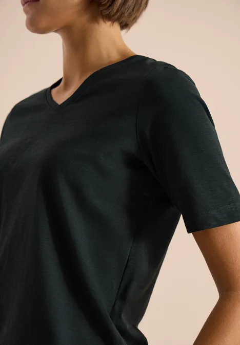 Basic Shirt met V-hals palm breeze green Zijaanzicht van een persoon in een zwart V-hals T-shirt met korte mouwen tegen een beige achtergrond.