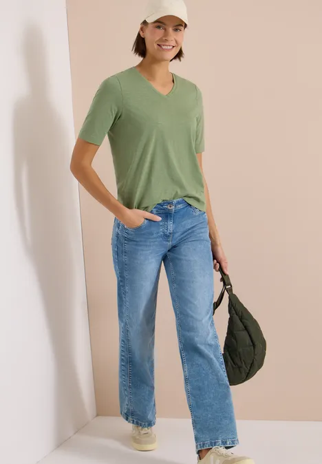 Kobieta w zielonym T-shircie, niebieskich jeansach, białych sneakersach, jasnej czapce i zielonej torbie.