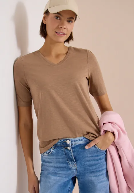 Frau in beigeem V-Ausschnitt-Shirt und blauen Jeans, hält einen rosa Blazer in der Hand.