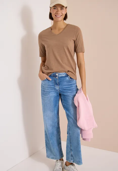 Frau in braunem T-Shirt und blauer Jeans hält rosa Pullover vor hellem Hintergrund.