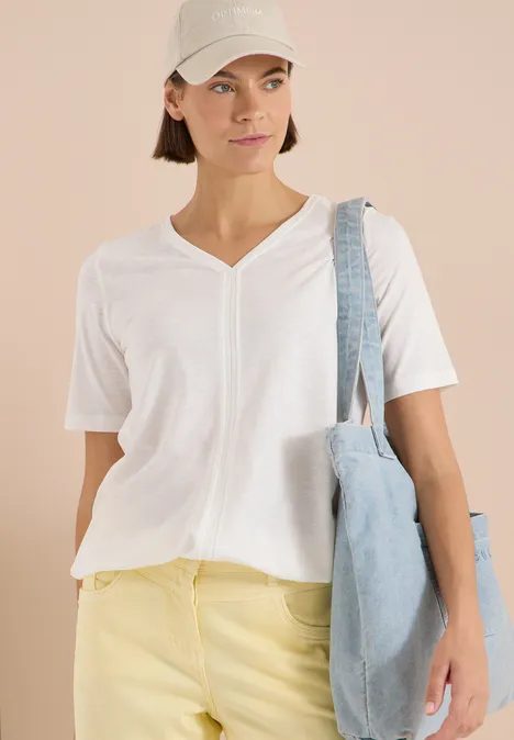 Femme porte un t-shirt blanc à col V, pantalon jaune, casquette blanche à l’arrière et sac bleu clair.