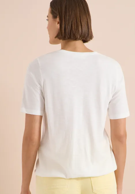 Femme de dos portant un t-shirt blanc simple et un pantalon jaune sur fond neutre.