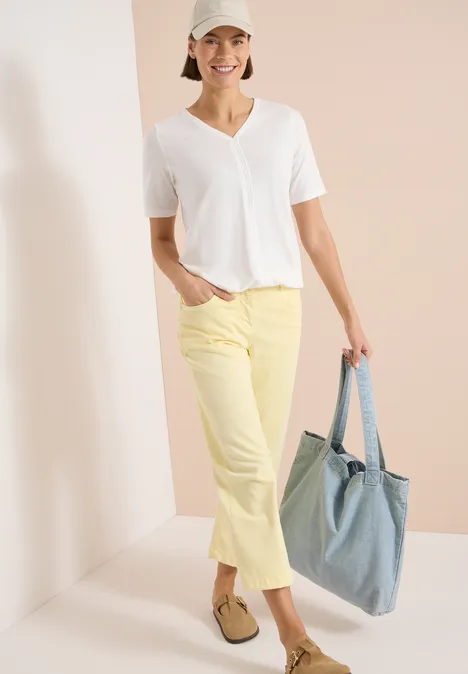Femme en T-shirt blanc col V, pantalon jaune court, mocassins et sac en jean devant un fond clair minimaliste.