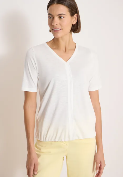 Une femme porte un t-shirt blanc à col V et un pantalon jaune sur fond neutre.