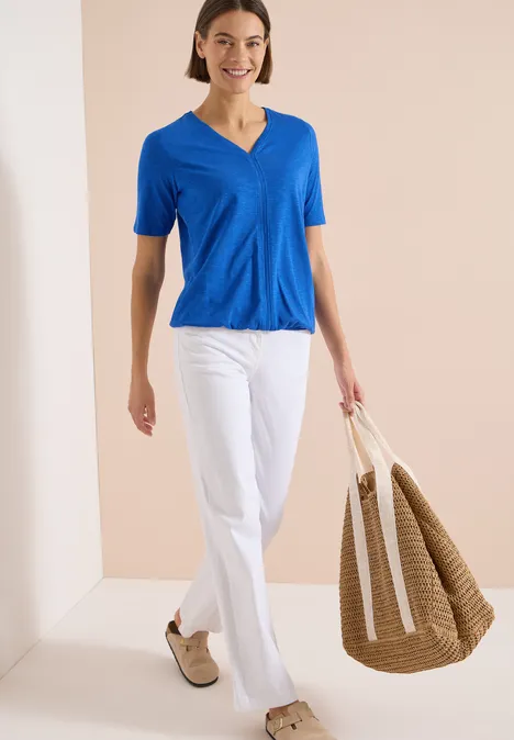 Kurzarm T-Shirt im Tunika-Look surf blue Frau in blauer V-Ausschnitt-Bluse, weißer Hose und beigen Schuhen trägt große geflochtene Tasche.