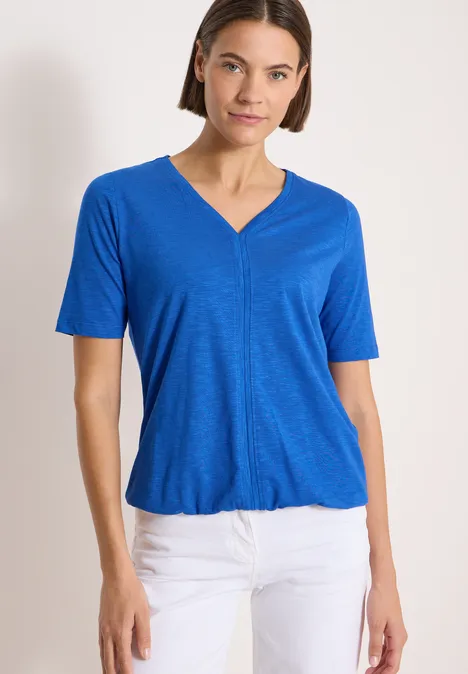 Kurzarm T-Shirt im Tunika-Look surf blue Frau trägt blaues T-Shirt mit V-Ausschnitt und weiße Hose, vor hellem Hintergrund.