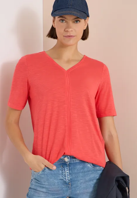 Personne vêtue d’un t-shirt rouge à col V et d’un jean bleu, tenant une veste sombre.