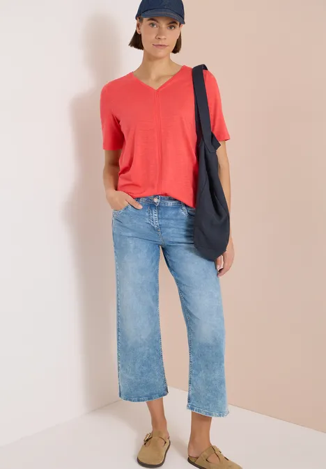 Personne en t-shirt rouge, jean large bleu, mules beiges et sac sombre sur fond clair.