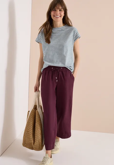 Een vrouw met een gestreepte top, paarse broek en een strooien tas.