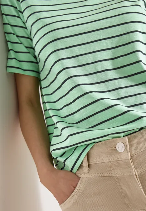 Person lehnt an Wand, trägt grünes T-Shirt mit schwarzen Streifen und bequeme beige Hose, eine Hand in der Tasche.