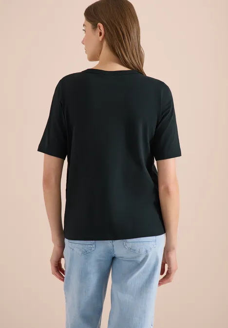 Frau von hinten in schwarzem T-Shirt und hellblauen Jeans vor hellbeigem Hintergrund.