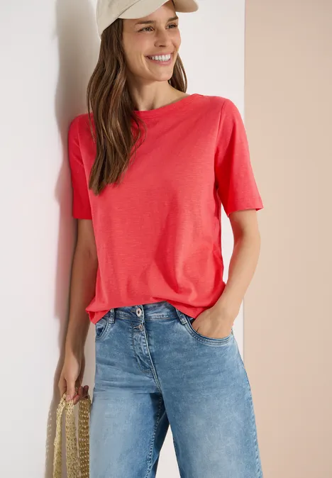 Kurzarm T-Shirt aus reiner Baumwolle coral Frau lehnt an weißer Wand, trägt rosa T-Shirt, blaue Jeans und hält eine geflochtene Tasche.