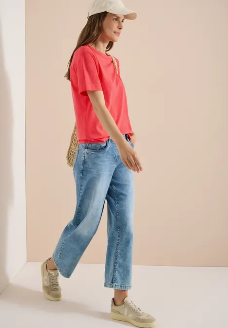Kurzarm T-Shirt aus reiner Baumwolle coral Frau im roten T-Shirt, blauen Jeans und Sneakers, seitlich stehend vor beigem Hintergrund.