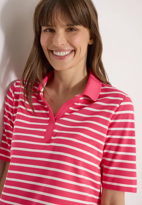 Mädchen trägt rosa-weiß gestreiftes Poloshirt mit kurzen Ärmeln vor neutralem Hintergrund.