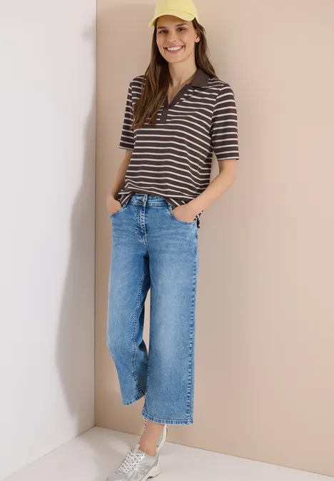 Person in gestreiftem Poloshirt, weiten Jeans und weißen Sneakern lehnt lässig an heller Wand.