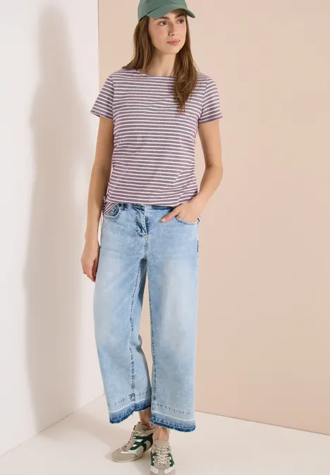 Person mit gestreiftem T-Shirt, weitem Jeans und Sneakern steht in einem hellen Eckzimmer.