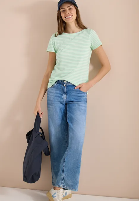 Persoon voor beige achtergrond met groen gestreept shirt, wijde jeans, witte sneakers en zwarte tas.